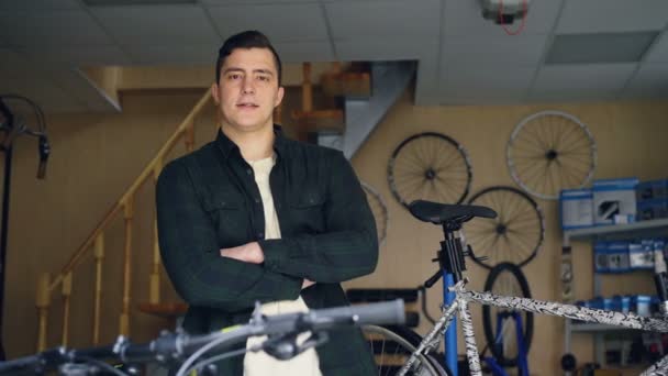 Retrato de joven guapo mecánico profesional de pie en taller de reparación de bicicletas con las manos cruzadas y mirando a la cámara. Bicicletas y repuestos son visibles . — Vídeo de stock