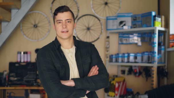 Retrato del propietario del taller de reparación de bicicletas de pie en su lugar de trabajo y mirando a la cámara. Repuestos para bicicletas, equipos de reparación y herramientas están en segundo plano . — Vídeo de stock