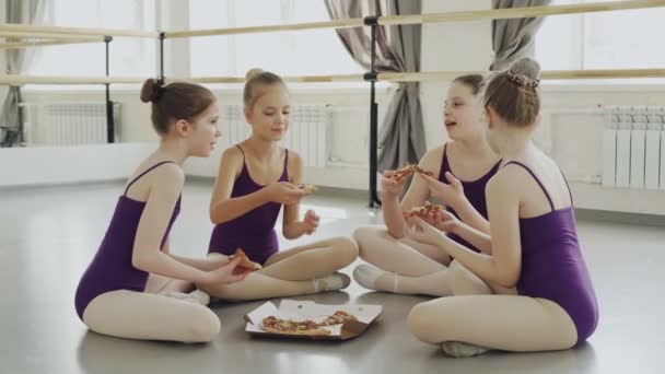 Glückliche Mädchen in hellen Trikots essen Pizza und unterhalten sich, während sie gemeinsam auf dem Boden des Ballettstudios sitzen. Leckeres Essen, Kommunikation und Kinderkonzept. — Stockvideo