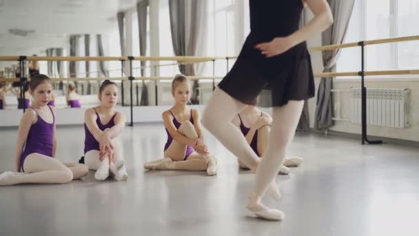 Профессиональная балерина-педагог танцует на цыпочках, демонстрируя движения своим маленьким ученикам, которые сидят на полу и смотрят на репетитора. . — стоковое видео