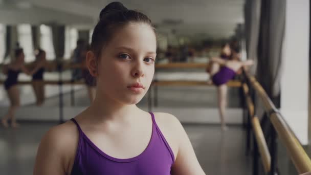 Ritratto ravvicinato di bambina mora in body in piedi in classe di danza classica e guardando la telecamera. Altri studenti praticano in background . — Video Stock