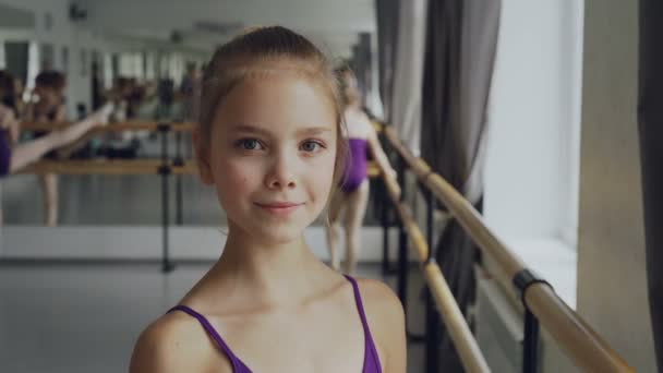 Ritratto ravvicinato di bella bambina in body in piedi in classe di danza classica, sorridente e guardando la macchina fotografica. Altri studenti stanno facendo esercizi in background . — Video Stock