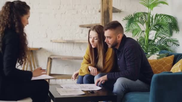 Adorables compradores de casas de pareja joven están mirando los papeles y hablando con el agente de bienes raíces luego sonriendo y besándose felizmente. Hermoso interior y muebles son visibles . — Vídeos de Stock