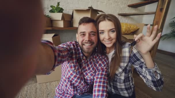 Фотография счастливой пары, делающей селфи с ключами от дома после покупки новой квартиры. Молодые люди держат камеру позируют и целуются с коробками на заднем плане . — стоковое видео