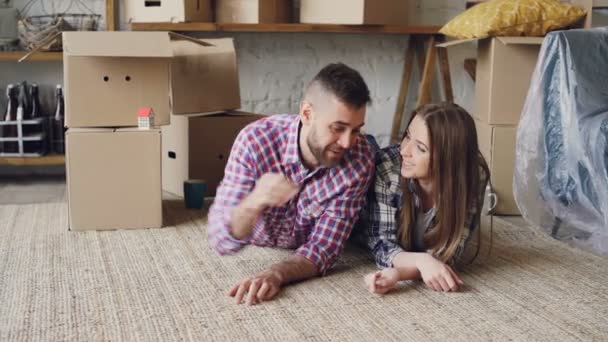 Lyckliga par liggande på golvet i nya hus, man ger nycklar till sin flickvän och kysser henne, de pratar och ler. Bostäder och relation koncept. — Stockvideo