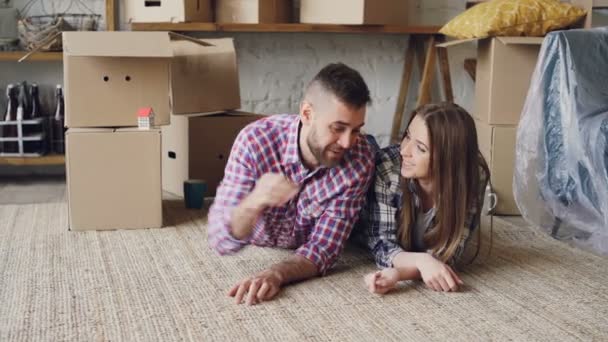 Pareja feliz está tirado en el suelo en la nueva casa, el hombre está dando las llaves de su novia y besarla, que están hablando y sonriendo. Concepto de vivienda y relación . — Vídeo de stock