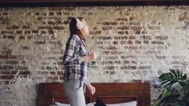 Mutlu neşeli kadın yatakta ev kablosuz kulaklık üzerinden müzik dinleme, dans ediyor. O rahat kıyafetler giyiyor, güzel ev iç görülür.