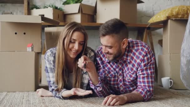 Chico barbudo alegre está dando la llave a su novia y besándola acostada en el piso de nuevo piso. Afecto, jóvenes, relación romántica y concepto de alojamiento . — Vídeos de Stock