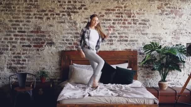 Bella studentessa in abiti casual sta ballando sul letto e cantando ascoltando musica con cuffie wireless. Giovani, tempo libero e stile di vita moderno . — Video Stock