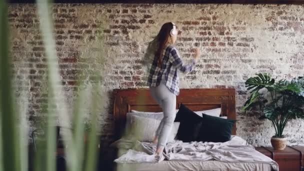 Schattig meisje is plezier dansen op bed in huis muziek beluisteren met koptelefoon springen en verhogen handen. Moderne levensstijl en gelukkige jonge mensen concept. — Stockvideo