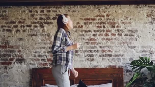 Szczęśliwy wesoły Kobieta tańczy się na łóżku w domu słuchania muzyki przez słuchawki bezprzewodowe. Ona ma na sobie ubranie, ładne wnętrze jest widoczny. — Wideo stockowe