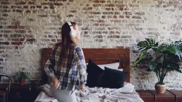 Attraktiv ung kvinna i casual kläder är dans på sovrum golvet och lyssnar på musik i hörlurar. Moderna loft stil lägenhet och växter är synliga. — Stockvideo