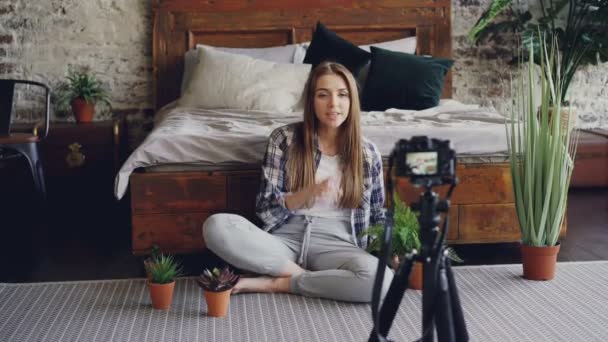 Jovem blogueiro sorridente em roupas casuais está segurando flores, falando e gravando blog de vídeo para vlog on-line sobre plantas de casa usando câmera no tripé . — Vídeo de Stock