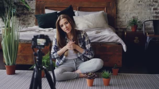 Chica bonita blogger popular está grabando video sobre flores de la casa con la cámara sentada en el piso cerca de la cama doble en un bonito dormitorio. Chica está hablando y sosteniendo la planta . — Vídeo de stock