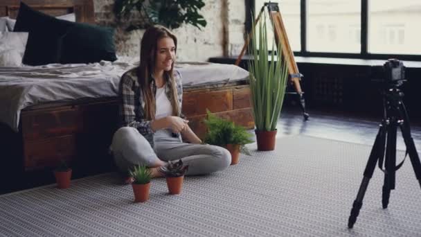 Joven blogger famosa está grabando video sobre flores de marihuana usando la cámara mientras está sentado en el piso del dormitorio cerca de la cama doble. Chica está hablando y mostrando plantas . — Vídeo de stock