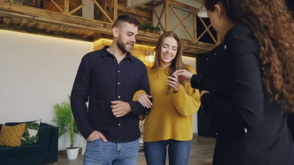 Movimento lento de corretor de imóveis amigável dando chaves para belos jovens compradores de casais de nova casa, os cônjuges felizes estão se beijando e olhando ao redor com prazer . — Vídeo de Stock