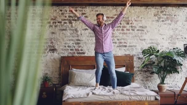 Lento movimiento de joven barbudo bailando en cama doble y escuchando música en auriculares. Apartamento de estilo loft moderno y plantas son visibles . — Vídeos de Stock