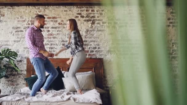 Cámara lenta de los amantes jóvenes felices bailando en la cama doble divirtiéndose en el dormitorio y riendo descuidadamente. Gente alegre, estilo de vida moderno y concepto de relación . — Vídeo de stock