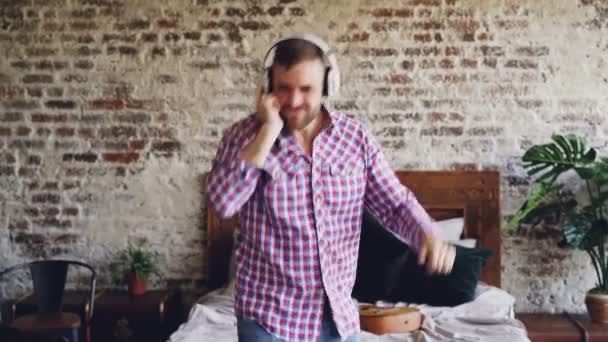 Neopatrný vousatý muž tančí a ukázal při poslechu hudby přes sluchátka v ložnici. Vzrušený chlap se baví ve volném čase relaxační. — Stock video