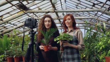 Önlükler içinde gülümseyen genç kadınlar bahçıvanlar konuşuyor ve video blog için fotoğraf makinesi tripod üzerinde kullanarak Bahçe hakkında online vlog kayıt çiçek tutuyorsun.