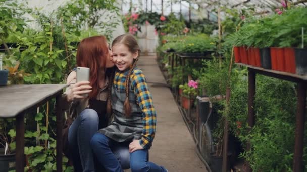 Young attractive worker of greenhouse and her cute emotional child are taking selfie hugging kissing and posing looking at smart phone camera. — Stock Video