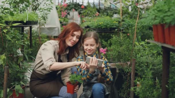 Sorridente giovane donna e il suo bambino carino stanno utilizzando smartphone, toccando lo schermo e ridendo all'interno della serra. Tecnologia moderna, famiglia felice e concetto di persone . — Video Stock