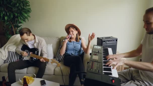 Glada vänner musiker praktiserande i huset, kvinnlig sångerska sjunger i mikrofonen och män spelar tangentbordet och gitarren tillsammans. — Stockvideo