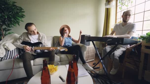 Dos chicos alegres músicos están tocando el teclado y la guitarra mientras su amiga vocalista está cantando sosteniendo el micrófono. Concepto de amistad y música . — Vídeo de stock