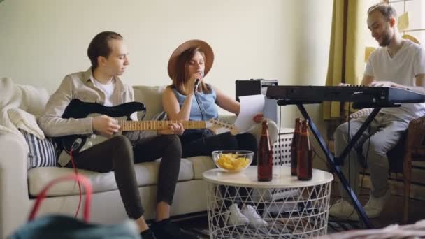 Die charmante Sängerin singt auf Papier mit Text, während ihre gutaussehenden männlichen Freunde Keyboard und Gitarre spielen. Tisch mit Flaschen und Snacks ist sichtbar. — Stockvideo