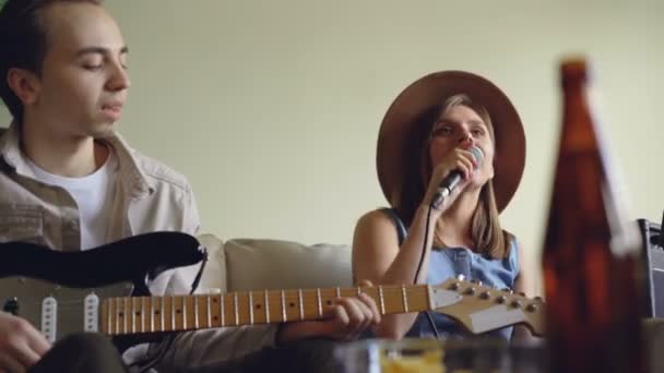 Linda joven está cantando y su guapo amigo guitarrista está tocando la guitarra eléctrica durante el ensayo en un bonito estudio de luz. Concepto de dúo creativo . — Vídeos de Stock