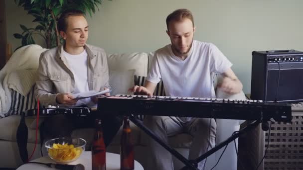 Jeune barbu clavier ajuste instrument de musique, puis jouer à regarder la partition et parler à son ami guitariste qui est assis sur le canapé en studio . — Video
