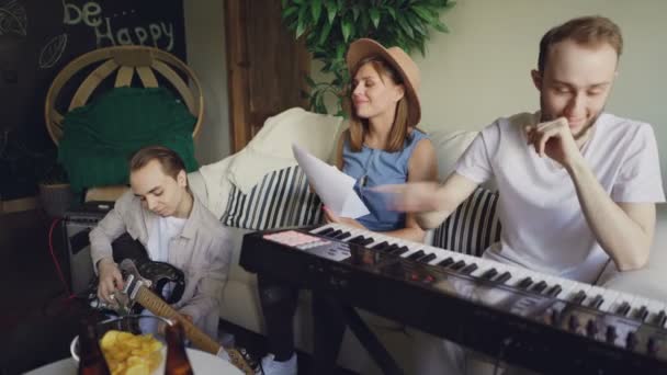 La banda musicale si sta esercitando a casa, il chitarrista è seduto sul pavimento e suona la chitarra, il cantante sorridente sta cantando, il tastierista sta ridendo e suonando la tastiera . — Video Stock