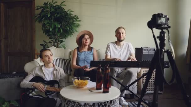 Jóvenes miembros de la banda popular están grabando video para blog en línea con cámara. Atractiva vocalista femenina está hablando sosteniendo micrófono, sus amigos están sonriendo . — Vídeos de Stock