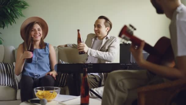 Amici allegri si divertono a parlare, cantare e bere birra durante le prove. Il chitarrista sta suonando, donna e uomo stanno battendo le bottiglie e ridendo . — Video Stock