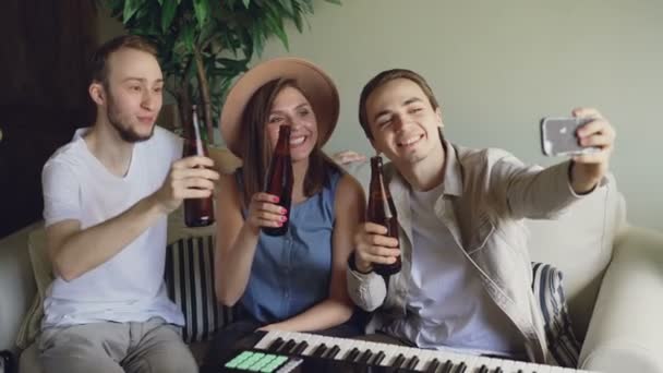 Les musiciens heureux prennent selfie avec des bouteilles de bière assis sur le canapé dans le home studio et tenant smartphone. Les jeunes posent, claquent des bouteilles et s'amusent . — Video