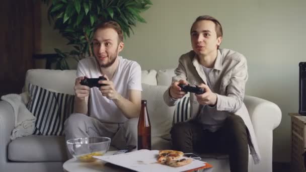 Två snygga unga killar spelar videogame holding joystickar sitter på soffan hemma. Känslomässiga män njuter av spelet, snacks och flaskor är synliga. — Stockvideo