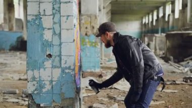 Grafiti sanatçısı sakallı adam parlak sprey boya sprey ile terk edilmiş binada ayağı üzerinde boyama. Kirli duvarlar ve kırık camlar ile boş endüstriyel yapı kadeh kaydırmak.