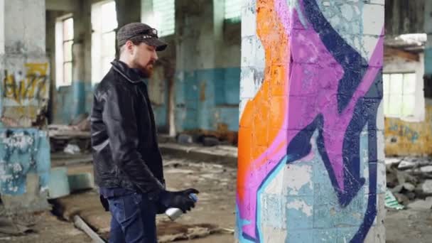 Knappe bebaarde man graffitikunstenaar is schilderen met verf binnen verlaten gebouw. Moderne straatkunst, jeugd subcultuur en creatieve mensen concept. — Stockvideo