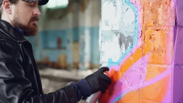 Vousatý chlap v neformálním oblečení je kresba abstraktní obraz barvou aerosolu na pilíř uvnitř staré budovy. Opuštěný dům, koncept subkultury a tvořivosti mládeže. — Stock video