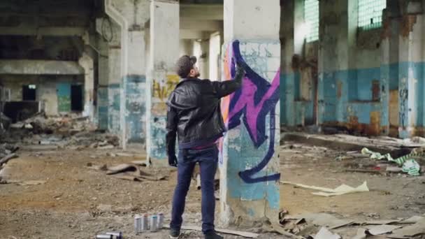 Graffiti pintor en ropa casual está dibujando con pintura en aerosol en la columna en el amplio edificio abandonado. Imágenes abstractas, arte moderno, gente creativa y concepto hipsters . — Vídeo de stock