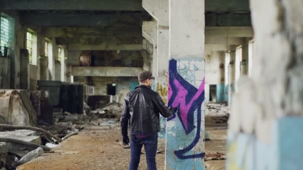 Male graffiti painter is creating abstract image with spray paint inside abandoned empty building. Old column is in foreground, dirty walls and windows in background. — Stock Video