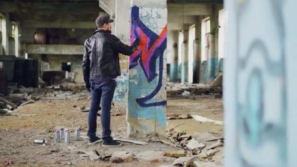 Szakállas fiatalember festészet graffiti belül régi ipari épületben segítségével aeroszolos festék, ő így absztrakt képek világos oszlop. A kreativitás és az emberek koncepció. — Stock videók