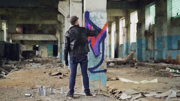 Back view of graffiti painter creating beautiful image with aerosol paint inside abandoned building. Artist is wearing blue jeans, black leather jacket, cap and gloves. — Stock Video