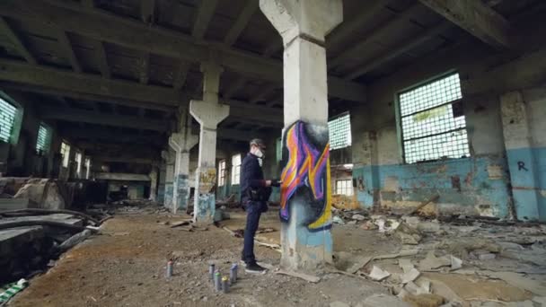 Graffiti artista en máscara protectora está pintando en la columna alta en el edificio industrial abandonado. Personas creativas, arte mural moderno y concepto de equipo de protección . — Vídeos de Stock