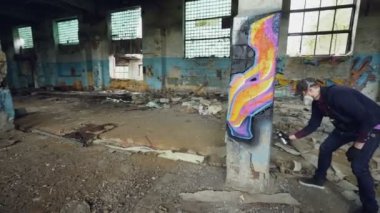 Maskeli grafiti sanatçısı ayağı boya sprey kullanarak büyük boş yapı olarak soyut görüntüleri çizim kadeh kaydırmak. Ressam casual giyim ve koruyucu eldiven giyiyor.