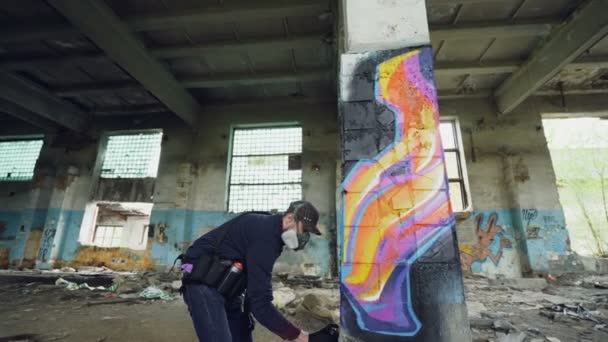 Bajo ángulo de disparo de hombre joven es la máscara de la cara de pintura de graffiti en la columna dentro de un edificio industrial vacío utilizando pintura en aerosol. Las paredes, ventanas y techos sucios dañados son visibles . — Vídeos de Stock