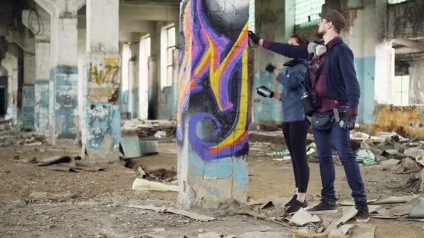 Bello barbuto uomo graffiti artista sta insegnando il suo amico donna a disegnare con vernice spray in piedi all'interno di vecchio edificio danneggiato e parlando in possesso di bombolette aerosol . — Video Stock