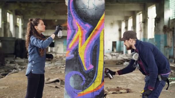Dos hábiles artistas de graffiti barbudo y atractiva joven están trabajando juntos en almacén abandonado decorando columna vieja con imagen abstracta . — Vídeo de stock