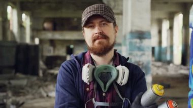 Yakın çekim yakışıklı sakallı adam grafiti sanatçısı kap, eldiven ve pespirator ve sprey boya tutan terk edilmiş binada ayakta portresi
