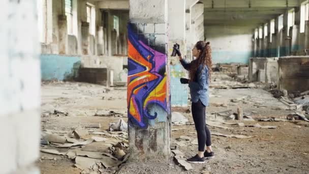 Chica bonita artista de graffiti está decorando vieja columna dañada dentro de un edificio industrial vacío con imágenes abstractas. Pintor moderno está utilizando aerosol pintura . — Vídeo de stock